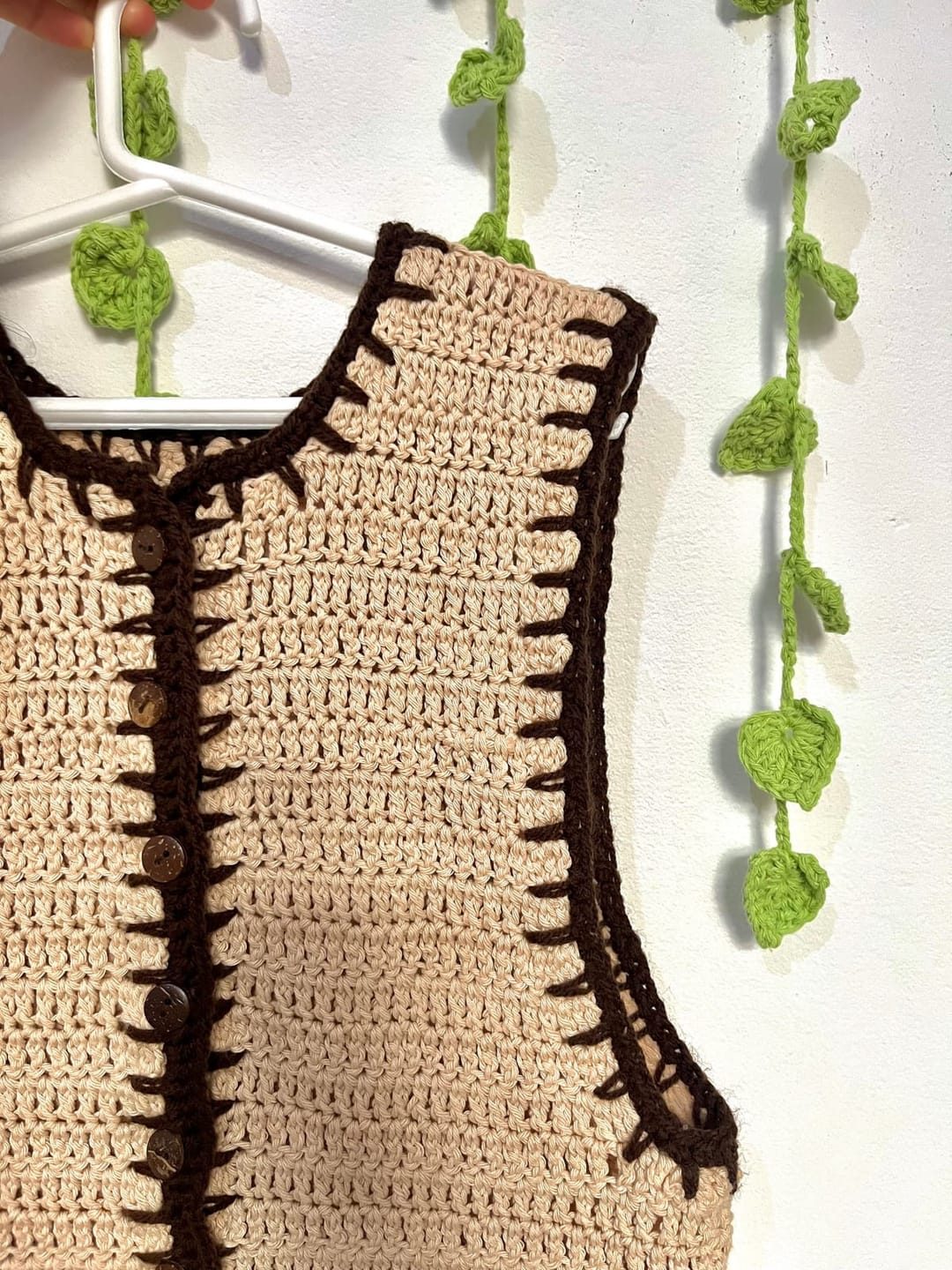 Chaleco tejido a crochet en algodón beige con borde marrón, colgado en una percha